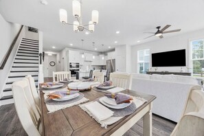 A modern kitchen with white cabinets, a dining area with a wooden table set for four, and a staircase with dark railings. - Golf-range View- Slps 11-Beach Shuttle-Grill- Pool (North Myrtle Beach)