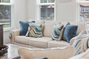 A beige sofa with multiple pillows, a glass coffee table, and a window with blinds. - Golf-range View- Slps 11-Beach Shuttle-Grill- Pool (North Myrtle Beach)