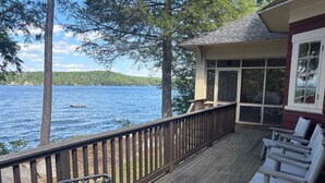 Una terraza de madera con una mesa y sillas de vidrio, con vistas a un lago con árboles y un edificio al fondo.