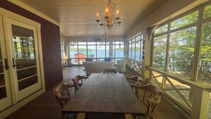Una terraza de madera con una mesa grande y sillas, una araña y ventanas amplias con vistas al agua.
