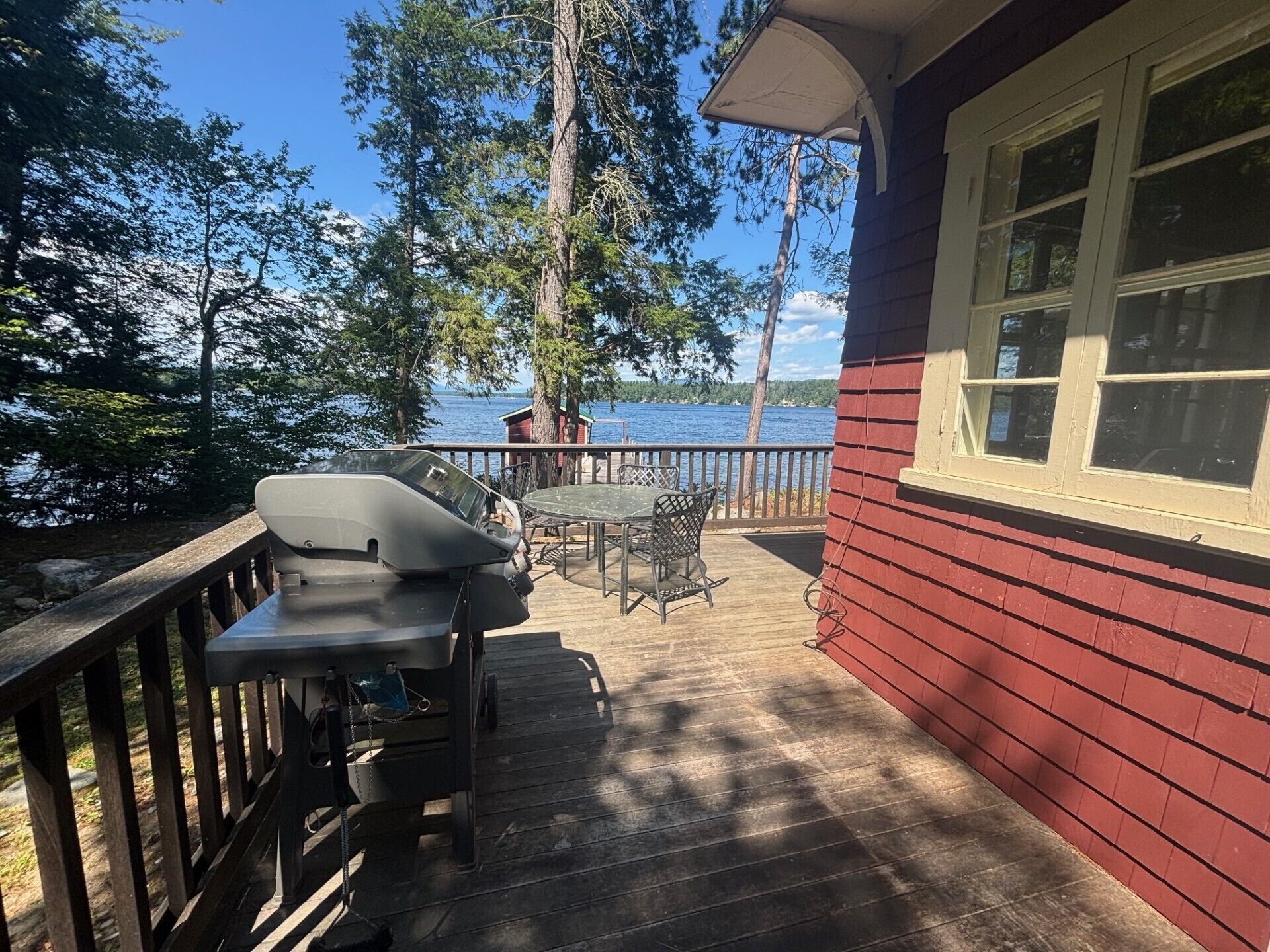 A wooden deck with a barbecue grill, a table, and chairs, overlooking a body of water and surrounded by trees.