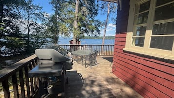 A wooden deck with a barbecue grill, a table, and chairs, overlooking a body of water and surrounded by trees.