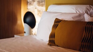 Una cama con almohadas blancas y color mostaza, un cabecero negro y una pared de piedra al fondo.