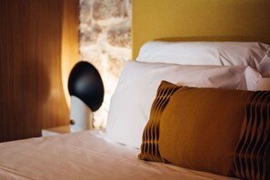 Una cama con almohadas blancas y color mostaza, un cabecero negro y una pared de piedra al fondo.