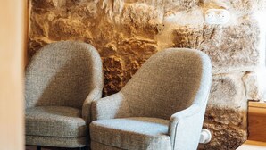 Twee grijze bekleden fauteuils tegen een geribbelde stenen muur, met een houten nachtkastje en een wit stopcontact.