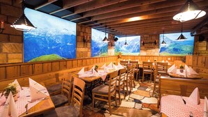 Un restaurante con mesas y sillas de madera, suelo de piedra y grandes ventanales con vistas a la montaña.