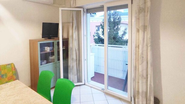 Ein Zimmer mit Esstisch, Stühlen, einem Fernseher und einer Glastür, die auf einen Balkon führt.