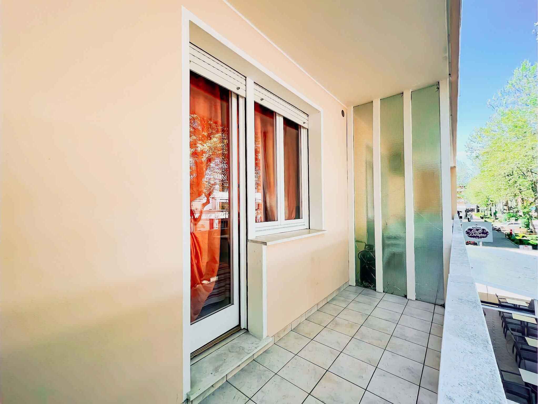 A balcony with tiled flooring and a view of a street with trees and a cafe.