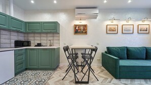 A compact kitchen with green cabinets, a microwave, and a dining table with chairs. There is a green sofa in the living area, and three framed pictures on the wall.