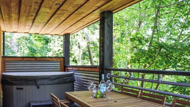 Une terrasse en bois avec un jacuzzi, une table et des chaises, et une vue sur une végétation luxuriante.