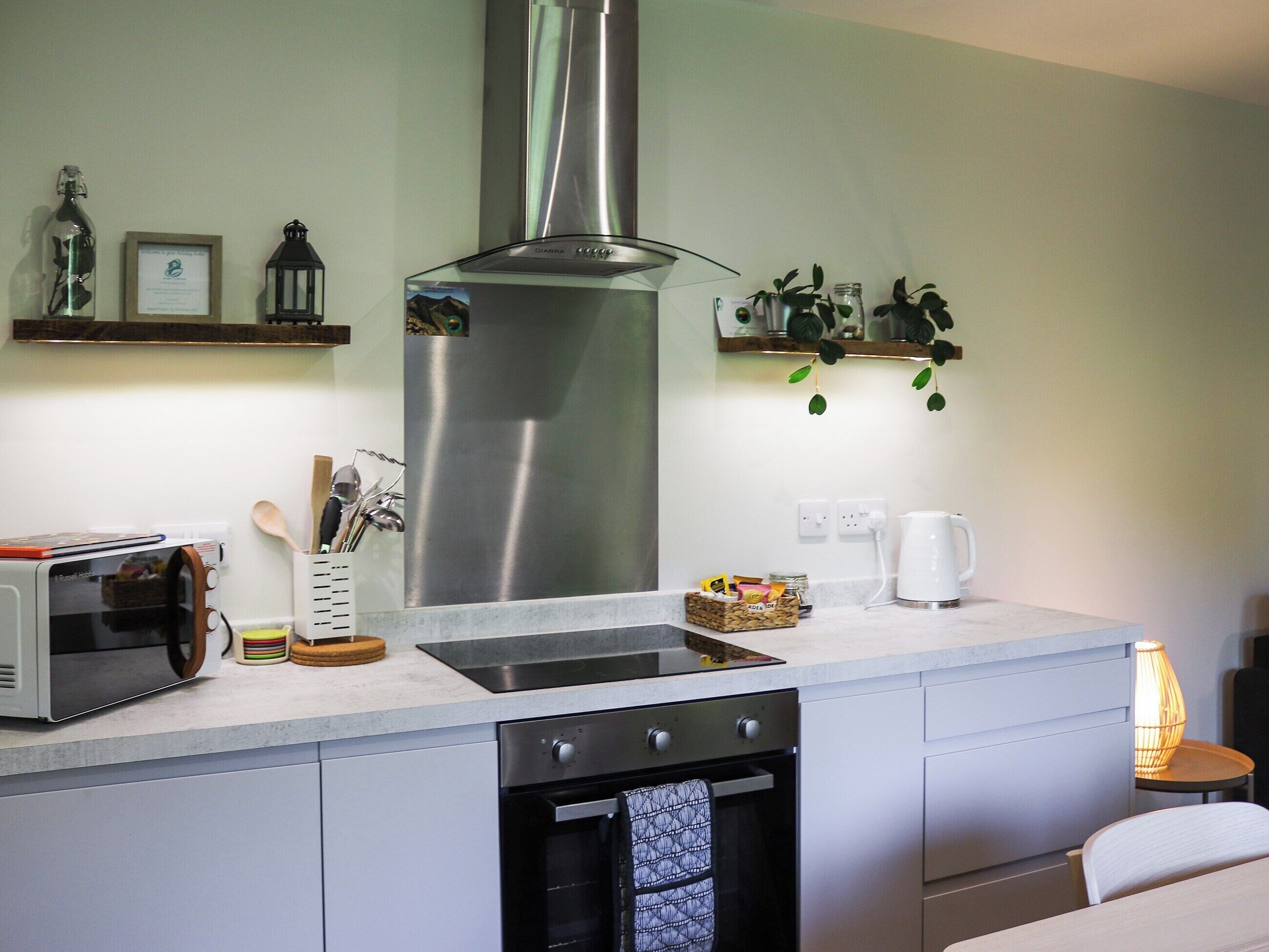 A compact kitchen with a stainless steel range hood, a built-in microwave, and a dishwasher. There are wooden utensils in a holder, a coffee maker, and a small shelf with plants.