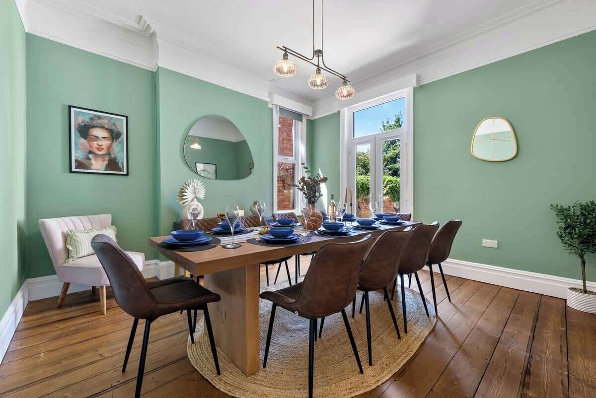 A dining area with a wooden table set for six, surrounded by chairs, a mirror, and a clock on the wall.