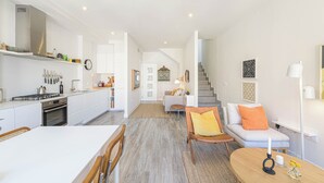A modern kitchen with white cabinetry, a built-in oven, and a stainless steel range hood. There is a dining area with a white table and chairs, and a living room with a sofa and a coffee table. - Solaga - Chinchilla (Málaga)