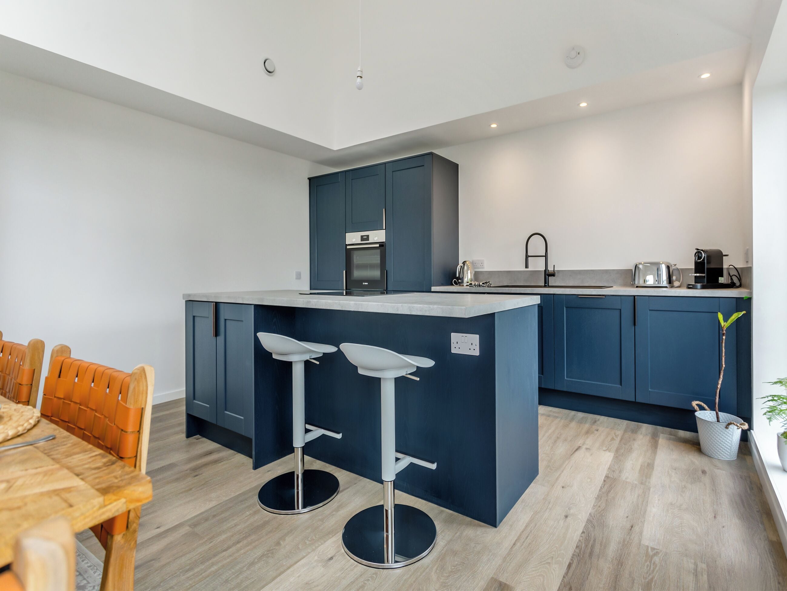 A modern kitchen with dark blue cabinets, a central island with bar stools, a wooden dining table, and a coffee machine.