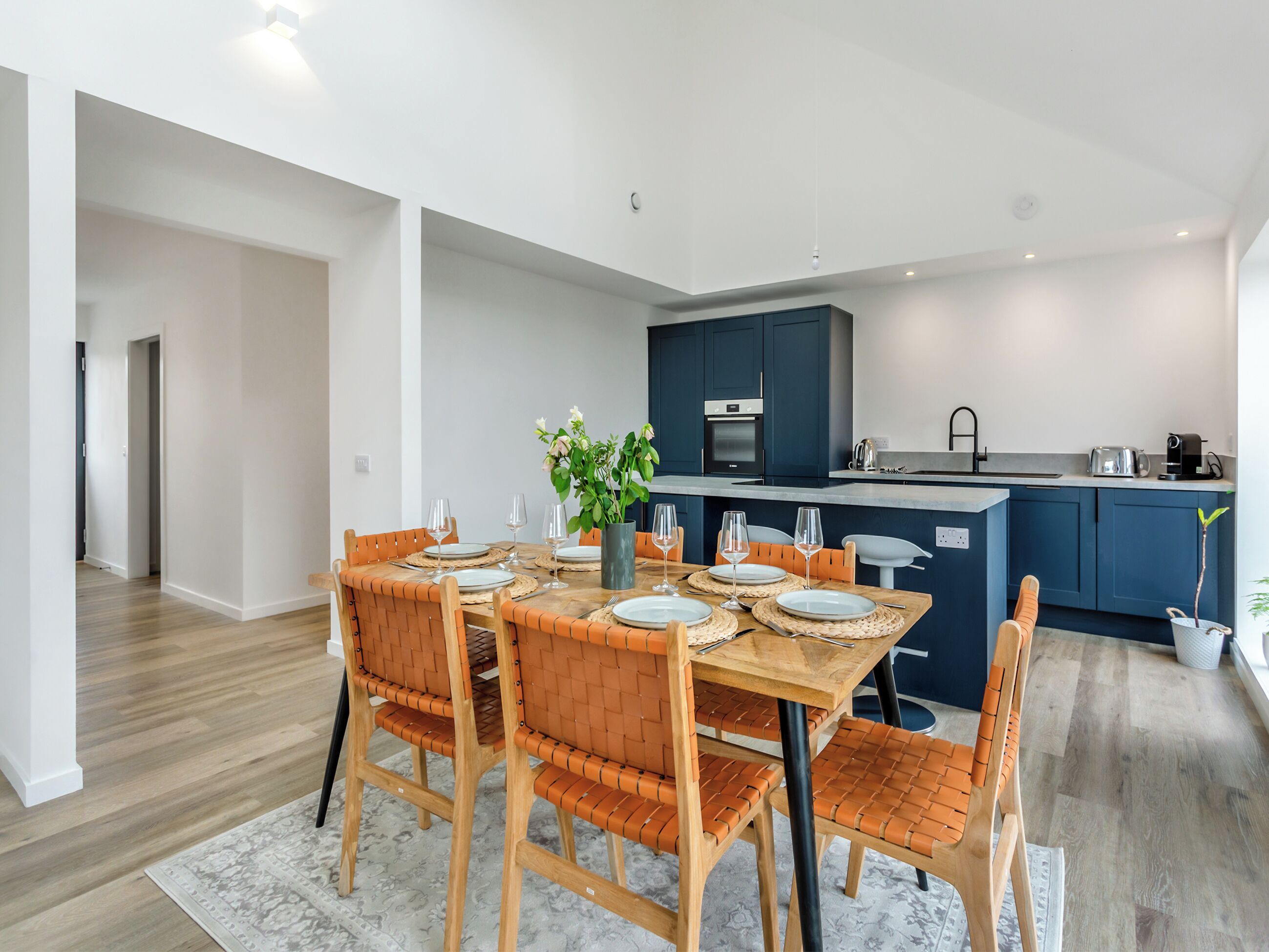 A modern kitchen with dark blue cabinets, a central island, and a dining area with a wooden table and orange chairs.