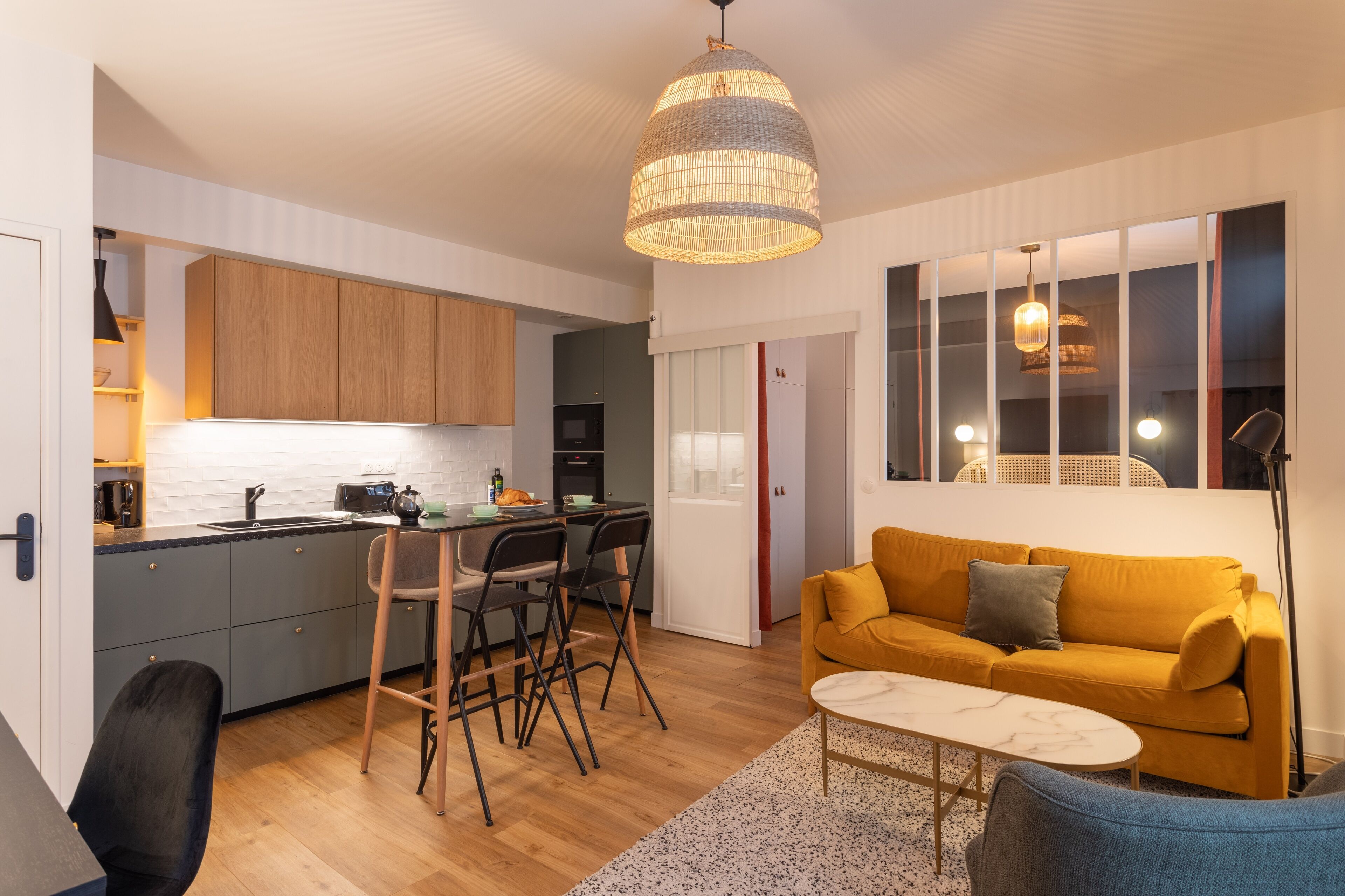A modern living room with a yellow sofa, a coffee table, and a dining area with chairs and a kitchen in the background.