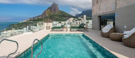 A rooftop pool with clear blue water, lounge chairs, and a view of a mountain and cityscape.