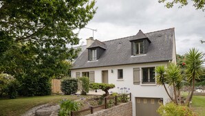 A two-story house with a stone staircase leading to a gated entrance, surrounded by lush greenery and a well-maintained garden. - Comtesse Lili - Maison proche de la mer, jardin (La Richardais)