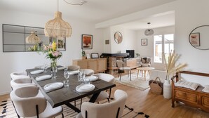 A dining area with a dark wooden table set for six, surrounded by white chairs. There is a woven pendant light above the table, and a vase with flowers on the table. - Comtesse Lili - Maison proche de la mer, jardin (La Richardais)