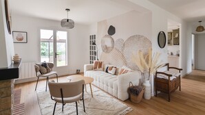 A modern living room with a sofa, a dining table, and chairs. There is a fireplace, a large window, and a kitchen area visible in the background. - Comtesse Lili - Maison proche de la mer, jardin (La Richardais)