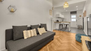 A modern living room with a grey sofa, wooden flooring, and a kitchen area in the background.