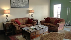 A living room with two sofas, a coffee table, and lamps. There is a framed picture on the wall and a window with a view. - Boarders Inn & Suites (Lamar)