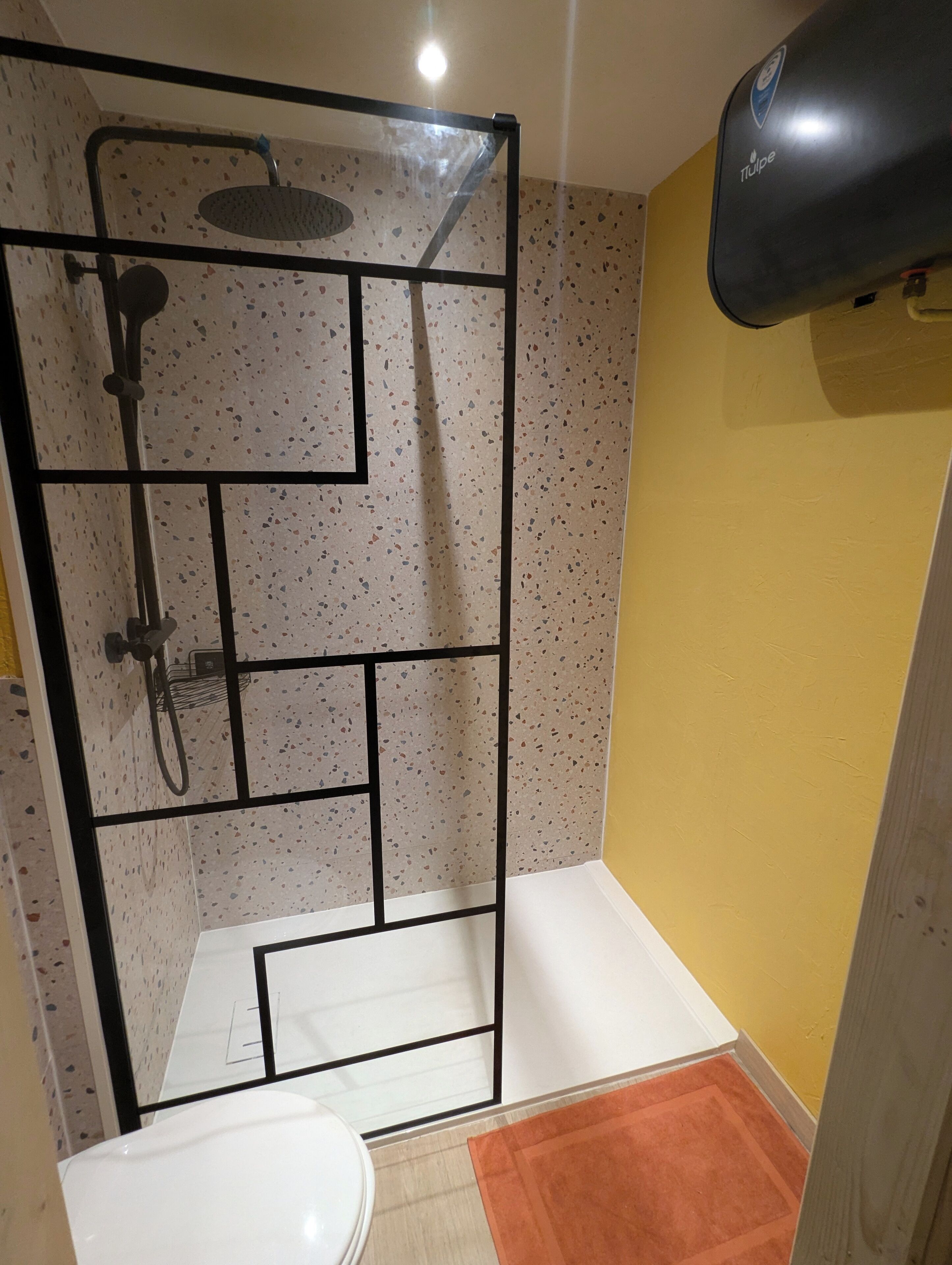 A shower enclosure with a rain showerhead and a handheld showerhead, a white toilet, and a yellow wall.