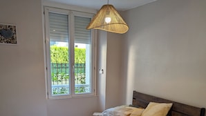 Een netjes opgemaakt bed met een bedovertrek met patroon en kussens, een hangende rotanlamp, een raam met blinds en een klein decoratief object aan de muur.