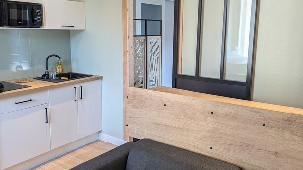 Een moderne keuken met witte kasten, een zwarte magnetron en een houten aanrecht. Er is een schuifdeur met uitzicht op een kamer met een bed en een raam.