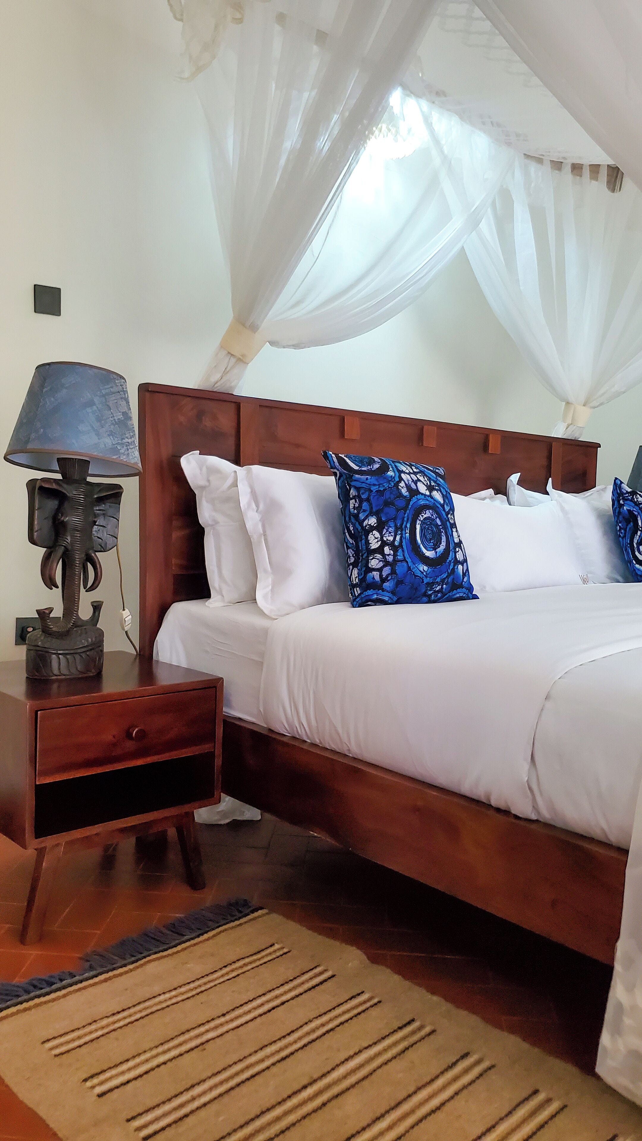 A bedroom with a wooden bed, white bedding, a blue patterned pillow, a nightstand with a lamp, and a mosquito net.