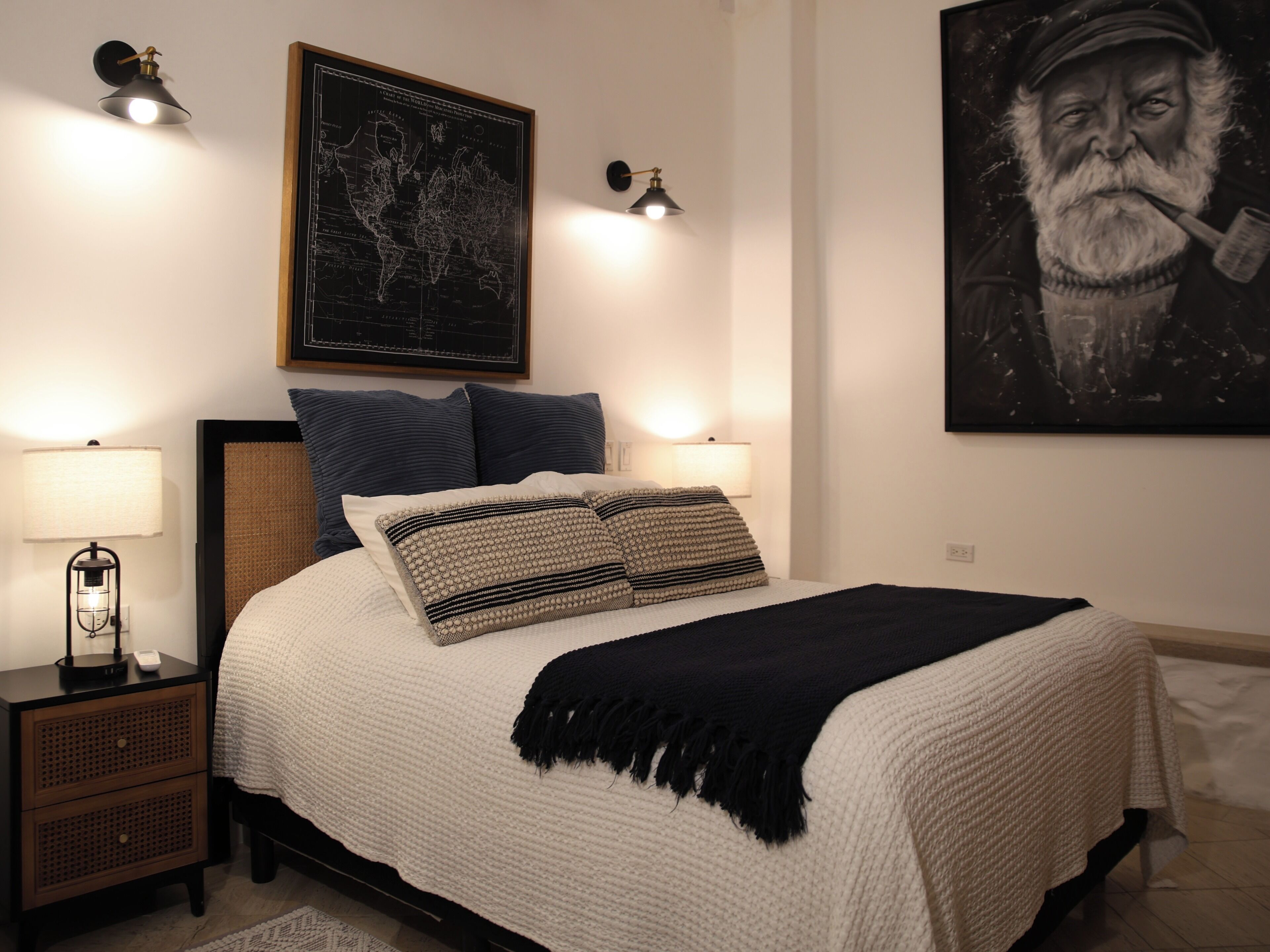 A neatly made bed with a white and beige blanket, two decorative pillows, a black throw, and a world map above it.