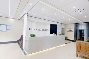 Zona de recepción de un hotel con mostrador de recepción identificado como 'CHUAN AO HOTEL', un banco de madera y una planta en maceta.