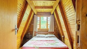 A cozy attic bedroom with a wooden sloped ceiling, a bed with a floral bedspread, and a window with curtains. - Chalé 3 Família (Angra dos Reis)