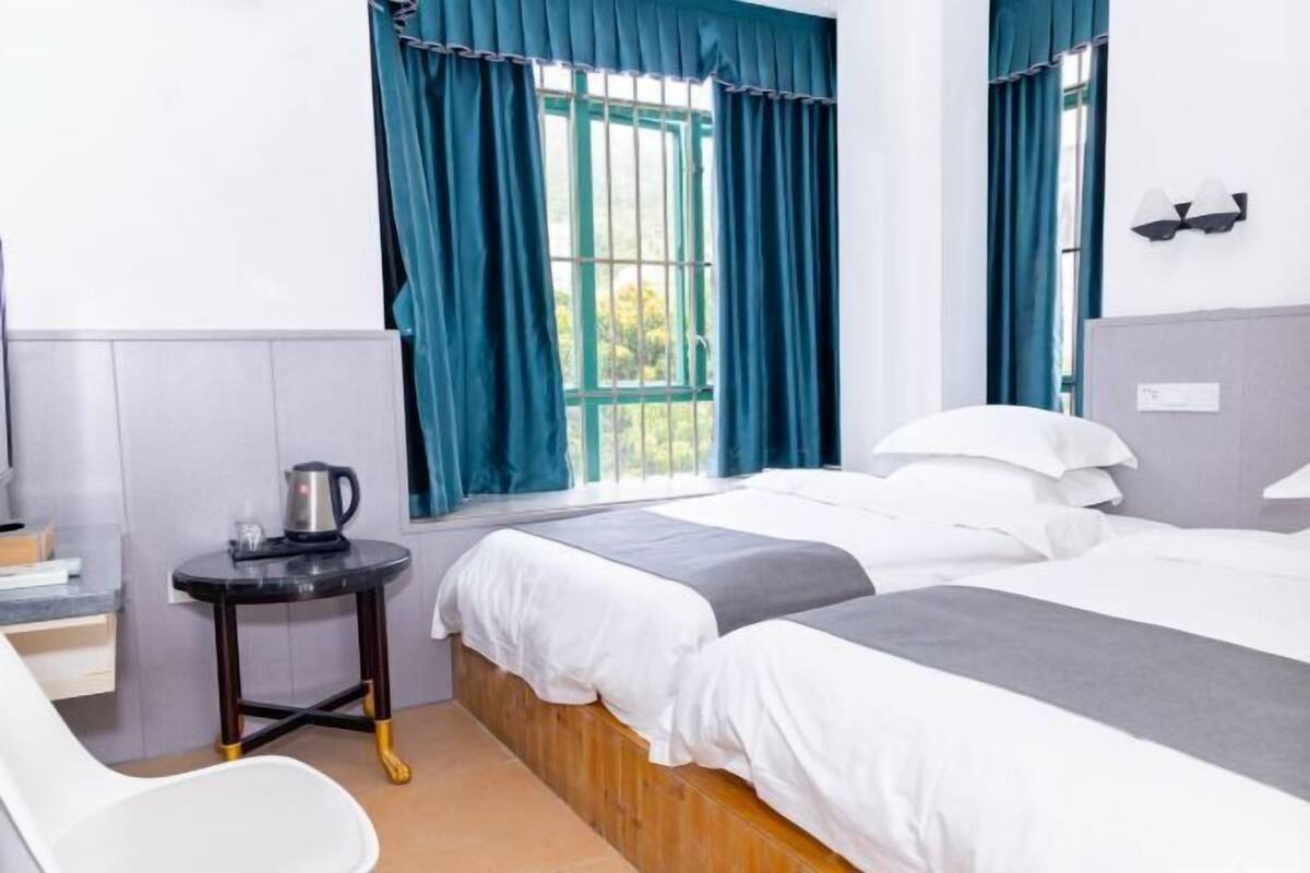 Habitación de hotel con dos camas, una mesita con hervidor, una silla y un ventanal con cortinas.