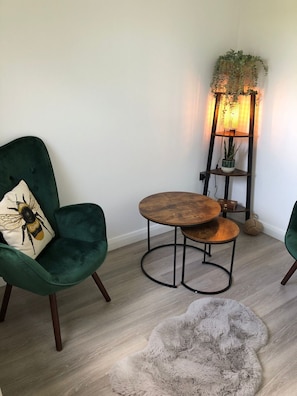 Una habitación de esquina con un sillón verde, una mesa redonda de madera, un tapete mullido y una lámpara de pie con plantas.