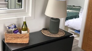 A black bedside table with a woven placemat, a lamp, a bottle of wine, and a basket containing snacks and a chocolate bar.