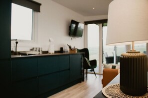 A modern kitchen with dark cabinetry, a flat-screen TV mounted above the counter, and a view of the outdoors through large windows.
