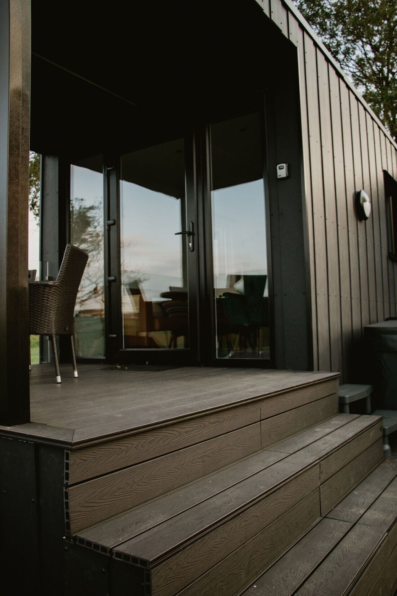 A wooden deck with a chair and a table, leading to a glass door entrance.