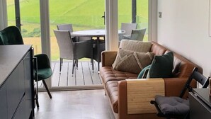 A modern living room with a large window, a leather sofa, a dining table with chairs, and a kitchen area.
