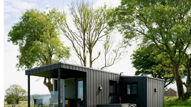 A modern, dark-colored cabin with a glass front, outdoor seating, and a hot tub.