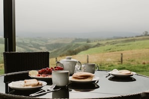 A dining table with plates of food, a teapot, and a cup, set against a view of rolling hills.