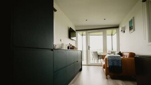 A modern kitchen with dark cabinets, a dining area with a brown sofa and a table, and a view through glass doors to an outdoor seating area.