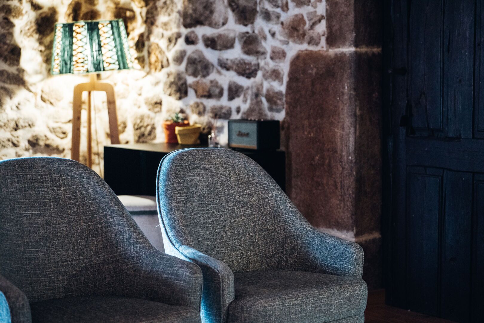 Una habitación con dos sillones grises, una mesa de madera, una lámpara y una pared de piedra.