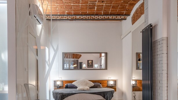 Un dormitorio moderno con techo de ladrillo, una cama con cabecera de madera, una mesita de noche y un espejo.