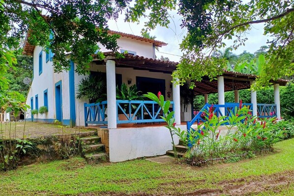A two-story building with a white exterior and blue accents, a covered porch with blue railings, and a garden area with various plants and a small water feature.