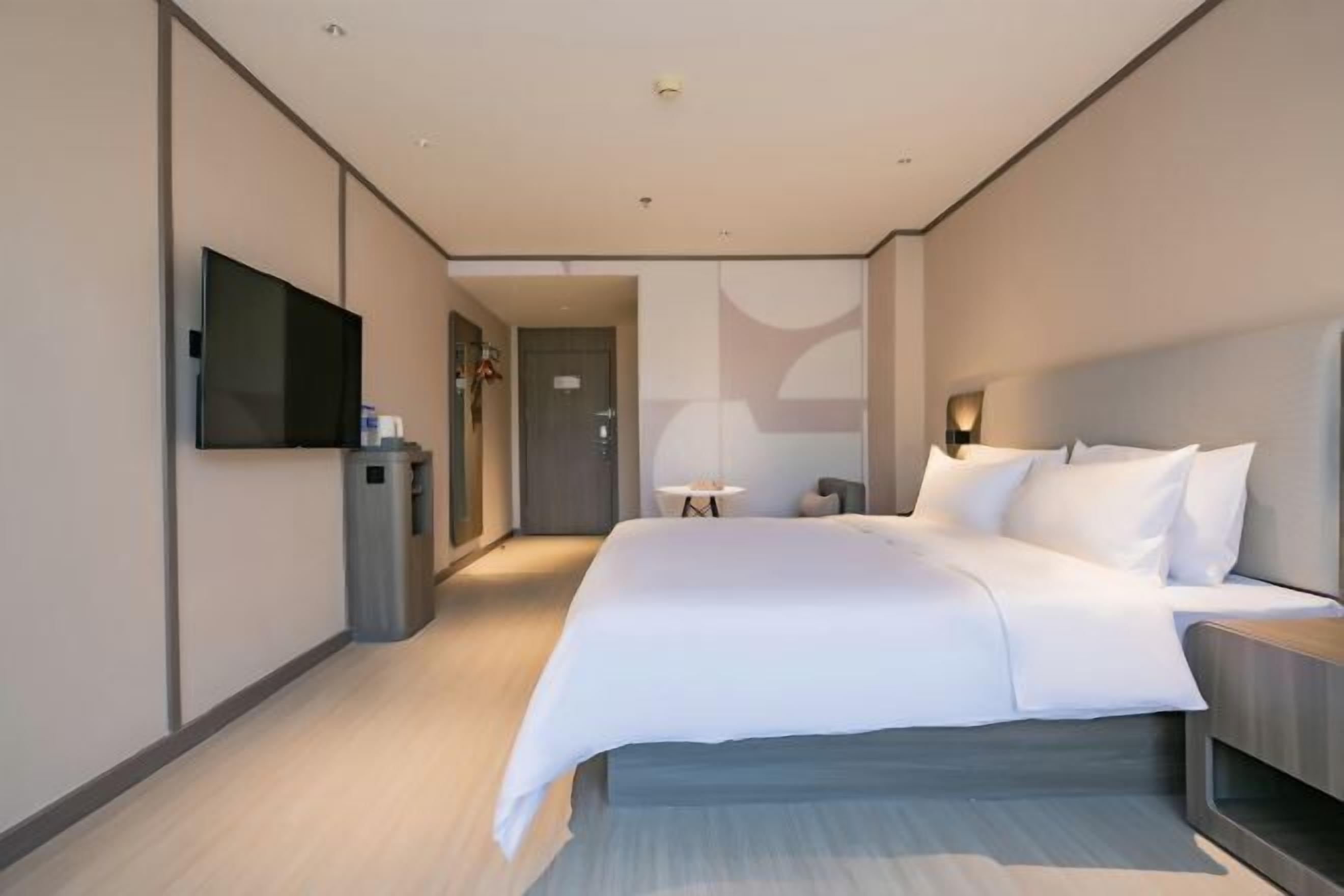 A modern hotel room with a large bed, a television mounted on the wall, a small table, and a door leading to another room.