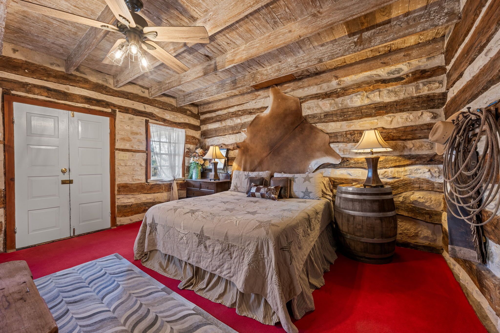 Une chambre de style chalet, avec un grand lit, un ventilateur de plafond en bois, un mur en pierre et une moquette rouge.