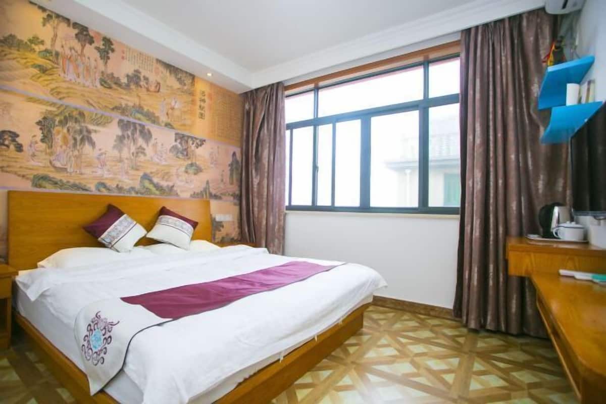 A hotel room with a large bed, a window with curtains, a wall with artwork, and a wooden desk with a cup and saucer.