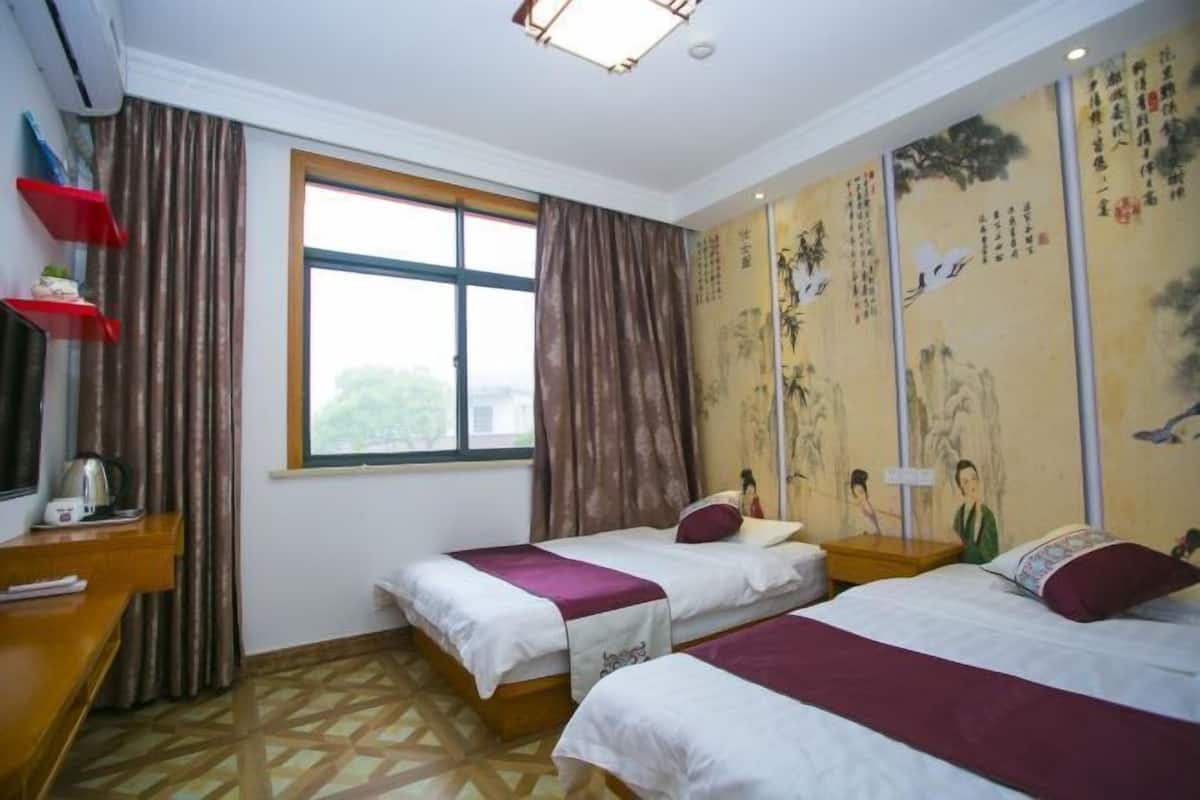 A hotel room with two beds, a window with curtains, a desk with a kettle, and traditional artwork on the walls.