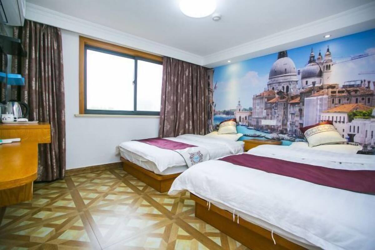 A hotel room with two beds, a window, a wall mural, a desk with a kettle, and patterned flooring.